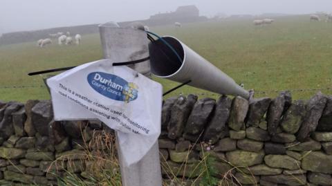 A toppled weather station. The metal pole of the weather station has been cut in half. The top half of the pole has fallen over a stone wall into a farmer's field. The field contains a herd of sheep. A small sign has been cable-tied to the pole explaining the device is a weather station and assists with public safety.