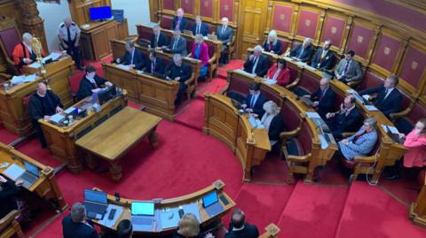 Members of the States of Jersey sat in the main chamber during a meeting. The room has red carpet, wooden desks and dark red leather seats.