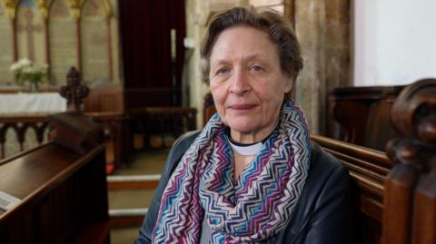 A woman wearing a clerical collar sits in a wooden church pew, looking towards the camera. The Reverend Sue Pulko is wearing a patterned scarf and a dark jacket. Behind her is the interior of a historic church, with carved wooden pews, stone walls and an altar.