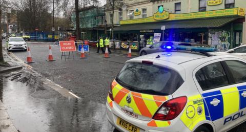 A police patrol car with yellow and blue bodywork is near the camera, while beyond it is a "road closed" sign, red and white traffic cones and police tape. Two officers in yellow high-viz jackets are seen by the cordon. In the background is a yellow-fronted shop called Malik's.