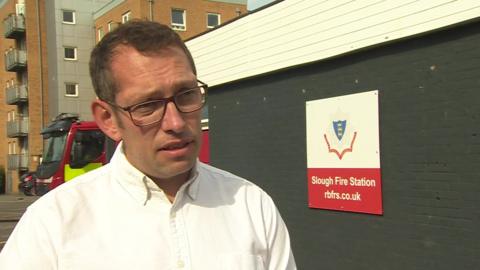 Steve Wright is wearing a white, buttoned shirt in front of a sign saying "Slough Fire Station". He is wearing dark-rimmed glasses and has short, dark hair. A fire engine and an apartment block are pictured behind him