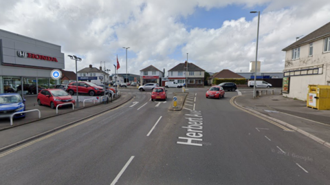 A Google street view screenshot of a road approaching a roundabout with a white building on the right and a large Honda garage on the left.