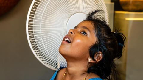 A little girl stands next to a whirring fan.