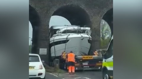 A large white boat on a trailer passing between two railway arches. A man in an orange florescent uniform is standing behind the trailer and a white car is travelling through the neighbouring arch 