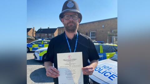 A police officer wearing a traditional tall police helmet and a black polo top holds up a white certificate outside with police cars parked behind him.