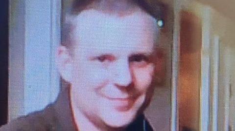 A man with brown eyes smiles at the camera. He is wearing a dark brown shirt. The image is a photo of a computer screen showing the picture of Mr Roberts.