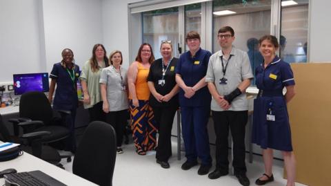 A line of people wearing various medical uniforms standing in an office with PCs, chairs and desks.   