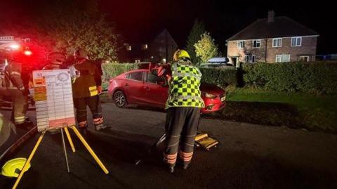 A firefighter standing in the road on a residential street at night. He is wearing a hi-vis vest and there is a large whiteboard being held up by a yellow tripod next to him. Other firefighters are also standing in the background. In front of them are two semi-detached houses.