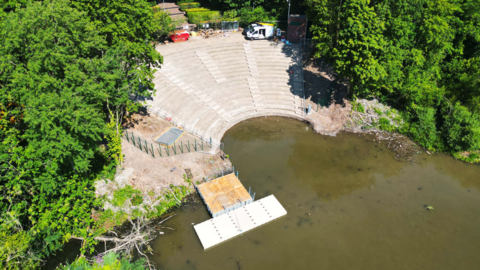An aerial photo of the amphitheatre show restored circular seating leading to a pool in the park. A pontoon sits on the left with some fencing still in place as work continues. Trees surround the site.