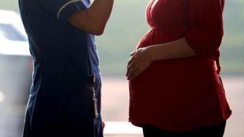 A nurse facing a pregnant woman. Their faces can't be seen. The nurse is wearing a blue uniform and the pregnant women is wearing a red dress.