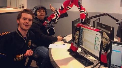 Webby, in a black top and flatcap and weaing headphones, in a BBC studio with a red, black and white Man Utd scarf