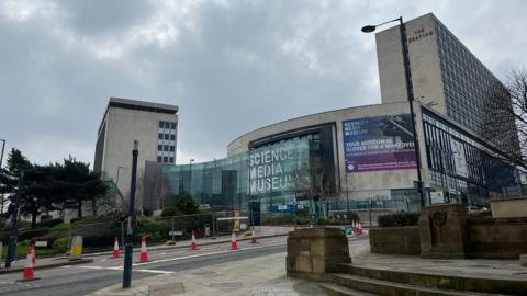 The front of the Science and Media Museum, which has a concrete facade behind a curved glass wall bearing the words Science + Media Museum in large white capital letters. On either side of the building are taller concrete tower blocks.