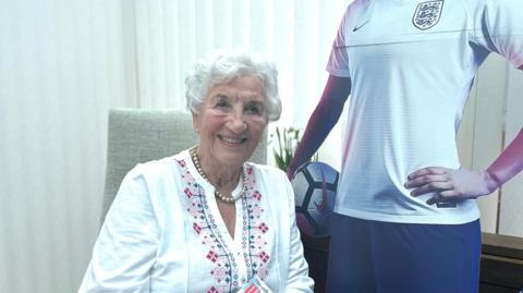 Gloria Stapleton sitting in a chair next to a life-size cardboard cut-out of her granddaughter Lucy Bronze. Mrs Stapleton is wearing a pearl necklace and a white blouse. She has short white hair and is smiling at the camera.