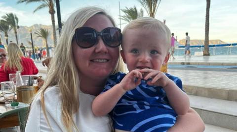 Hayley holds her baby Oliver in her left arm at a beachside restaurant. Oliver wears a navy blue striped top and has blond hair. Hayley wears black sunglasses and a white t-shirt. She also has blonde hair.