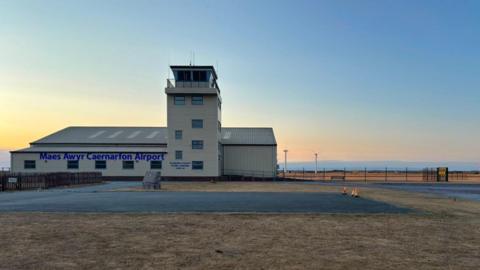 A small cream building airport. It has a watchtower and surrounded by green grass and fields. It also has car parks all around.