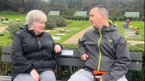 Two people are sat on one of the benches in Saltash. The lady on the left is wearing a black coat and the man on the right is wearing a grey coat.