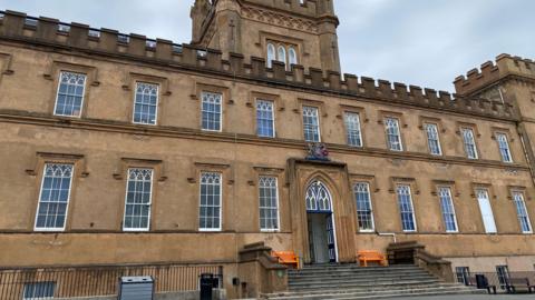 The image shows the exterior of Elizabeth College. It is a grand, two-storey stone building which has crenellations along the roof line and rectangular windows. There are also two towers. There are steps leading up to the front door which is open.