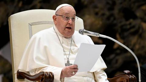 The Pope, speaking into a microphone while seated and wearing white robes.