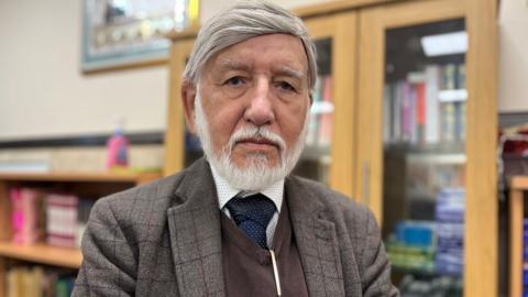 Abdul, a man with short white hair and white beard. He is wearing a checked jacket and a blue tie.