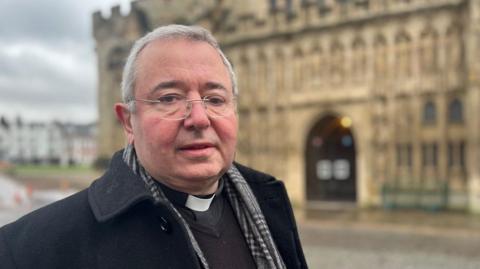 The Dean of Exeter, the Very Reverend Jonathan Greener, wearing a scarf and a black coat and standing in Cathedral Green with the cathedral in a blurred background.