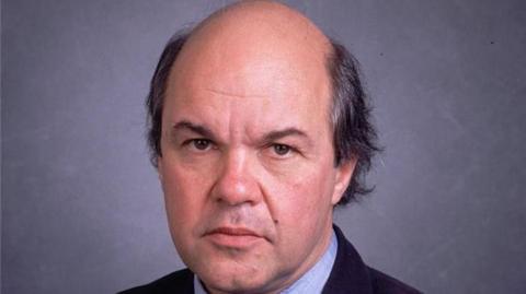 Portrait photo of a man with balding black hair. He is looking at the camera against a grey background, and wearing a black suit and blue shirt.