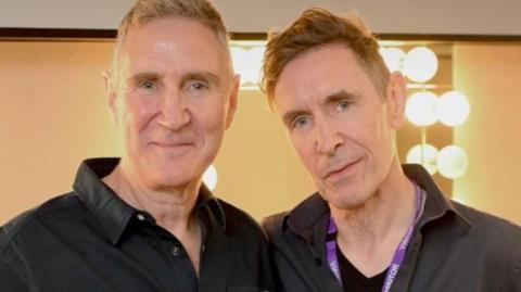 Mark and Paul stand next to each other with a make-mirror behind them. They're both wearing black shirts and smiling at the camera. 