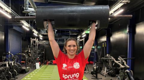Woman wearing red T shirt and weight lifter belt stands in a gym on a green baize runway with exercise machines on both sides. She is lifting a weight above her head