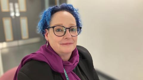 Fran, a woman with blue hair and wearing a purple scarf, smiles for the camera.