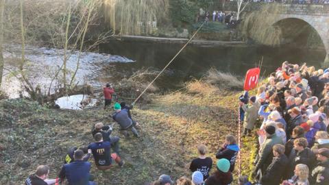 A group of people on a grassy slope. A long rope extends from this group toward the river and the group standing on the opposite river bank.
To the right side of the image, a large crowd of spectators is gathered behind a barrier, watching the activity. Many of these spectators are dressed in winter clothing, including coats and hats. A bright red life preserver is mounted on a post near the riverbank for safety. In the background, across the river, more people can be seen standing along the stone bridge, which has an arch visible on the right side.