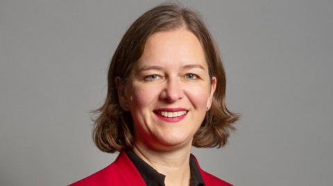 Fleur is standing against a professional looking grey wall, smiling.
The photo is cropped from shoulder height upwards.
She has blue eyes and light brown hair to her shoulders. 
She is wearing red lipstick and silver stud earrings.
She is wearing a black v neck blouse under a red collarless blazer. 