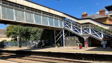 People waiting at the platform at Stroud Station, with the Grade II listed footbridge shown over the train tracks