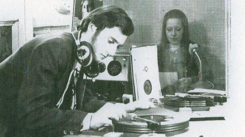 The photo is black and white, featuring a man leaning over to put a record on in a radio studio. He has dark hair, sideburns, headphones around his neck and is wearing a black suit jacket. There is a woman sitting in a booth behind him, reading a script behind a microphone.