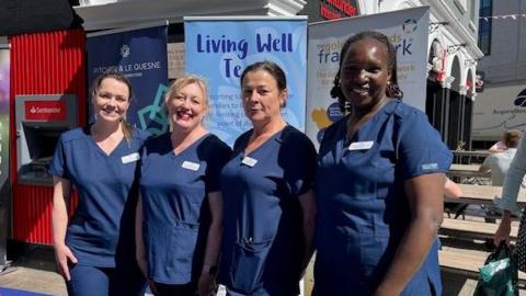 Four female nurses dressed in blue scrubs standing in front of banners advertising the Living Well Team.
