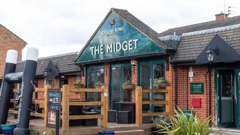 The front of The Midget pub in Abingdon. It is red brick with an arch like entrance. The pub's name is in large white letters on a green background