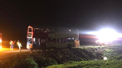 A red double-decker bus next to a ditch at night-time. It is upright but its roof has been crushed in. It is being lit up by emergency service's lights. 