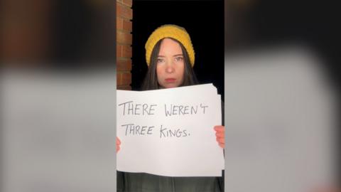 A woman in a yellow beanie hat holds up a piece of paper with the words 'There weren't three kings'