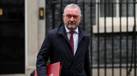 Steve Reed in a dark overcoat, white shirt and red tie walks outside a government building, carrying a large red document folder under one arm. The background features stone walls, a doorway and black metal railings. The photograph is taken at street level with the person in sharp focus and the building softly blurred behind.