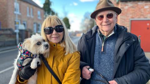 A couple in their 70s, in Keyworth. Jackie, left, is a white woman wearing a yellow padded coat with blonde hair and large black sunglasses. She is carrying a small dog wearing a multi-coloured harness. Paul is wearing a black hooded coat with a blue fleece underneath and a brown hat. 