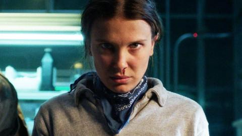 Stranger Things character Eleven, a teenager girl, is stood glaring directly at the camera. She is wearing a grey jumper with a navy, patterned neckerchief visible around her neck. A drip of blood is running from her nose towards her lip. Her head is lowered with her dark hair tied behind her head. Behind her, metallic shelves are visible, lit by neon lights and stacked with what appears to be bottles.