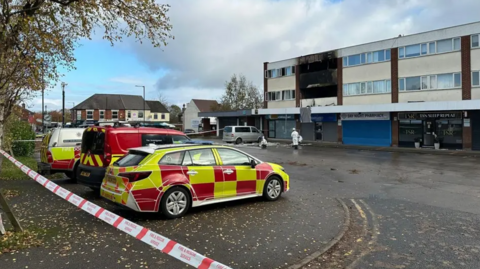 Police cars and ambulance vehicles are parked by the small block of flats with one flat clearly fire damaged and blackened with windows and their framework blown out