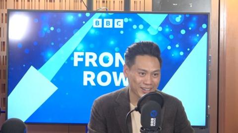 Jon M Chu speaking into a microphone in front of a large screen which says BBC Front Row