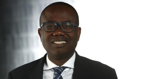 Kwesi Nyantakyi wears black-framed glasses, a white shirt, blue pattered tie and a dark suit jacket as he looks into the camera.