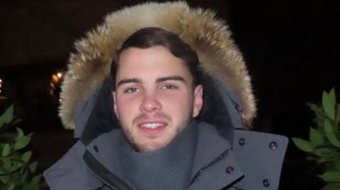 A young man with short black hair and facial hair. He is stood outside a night smiling at the camera. He is wearing a grey parka coat with the hood up