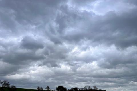 Dark, heavy storm clouds blanket the sky over a rural landscape, with a low horizon of fields and scattered trees 
