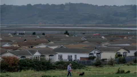 A wide shot of a series of white houses with grey roofs. In front of the houses there is green land and a man wearing a purple coat walking a black dog.