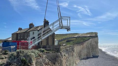 Metal steps being hoisted over a cliff, with buildings and the beach in the background.