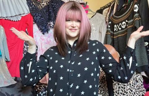 Caroline in her spare room, full of charity shop items. She is holing her arms out, wearing a pink and brown wig, with a black top with stars on, with clothing all around her. 