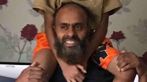 Masrob Ali pictured with a child on his shoulders. He wears a t-shirt and is smiling. He appears to be standing in a living room.