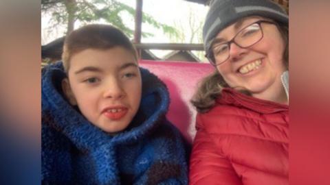A selfie of a mum, who is wearing a red coat, grey beanie hat and black glasses, and her son, who is wearing a blue fleecy hoodie. They are sat at the back of a land train in a woodland setting.