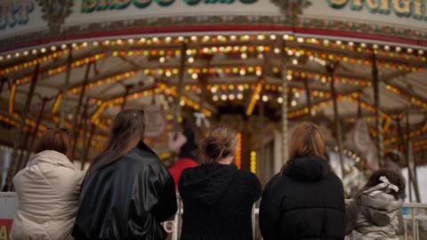 Four women stand in a row, with their backs to the camera, looking at a funfair carousel going round